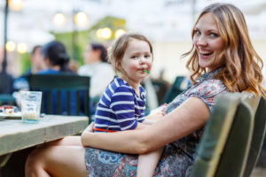 Little kid girl and her beautiful mother relaxing together with her little daughter, adorable toddler girl, in summer outdoors cafe drinking coffee on warm summer evening